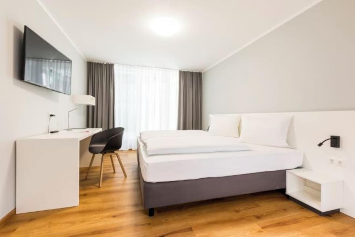 Una habitación de hotel moderna con cama, escritorio, silla y televisor en la pared.