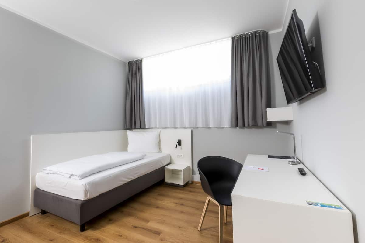 Une chambre d’hôtel moderne équipée d’un lit simple, d’un bureau, d’une chaise, d’un téléviseur à écran plat fixé au mur et d’une fenêtre avec des rideaux.