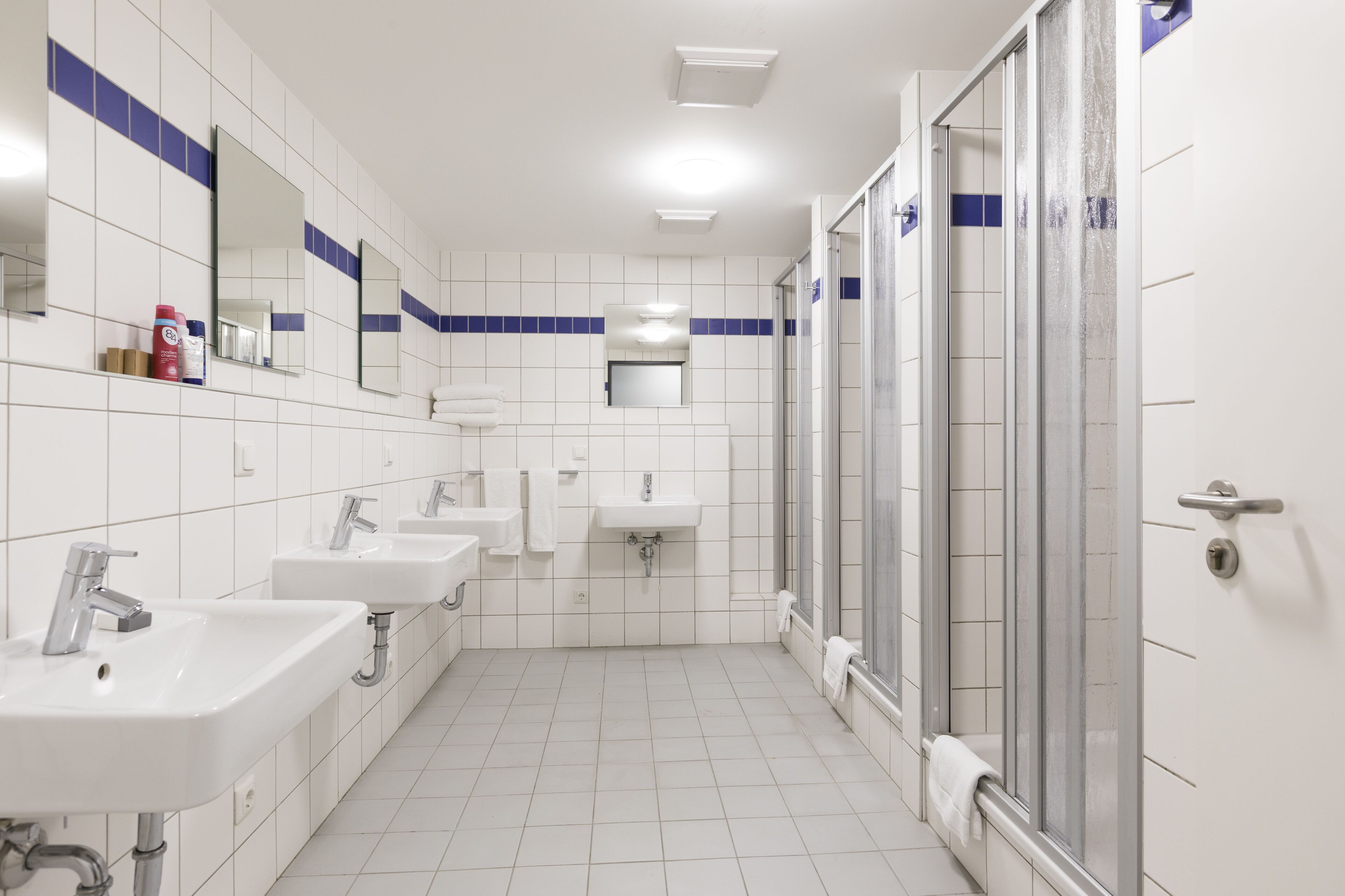 Un baño moderno con azulejos blancos, varios lavamanos, cabina de ducha con mampara de vidrio y un espejo sobre los lavamanos.