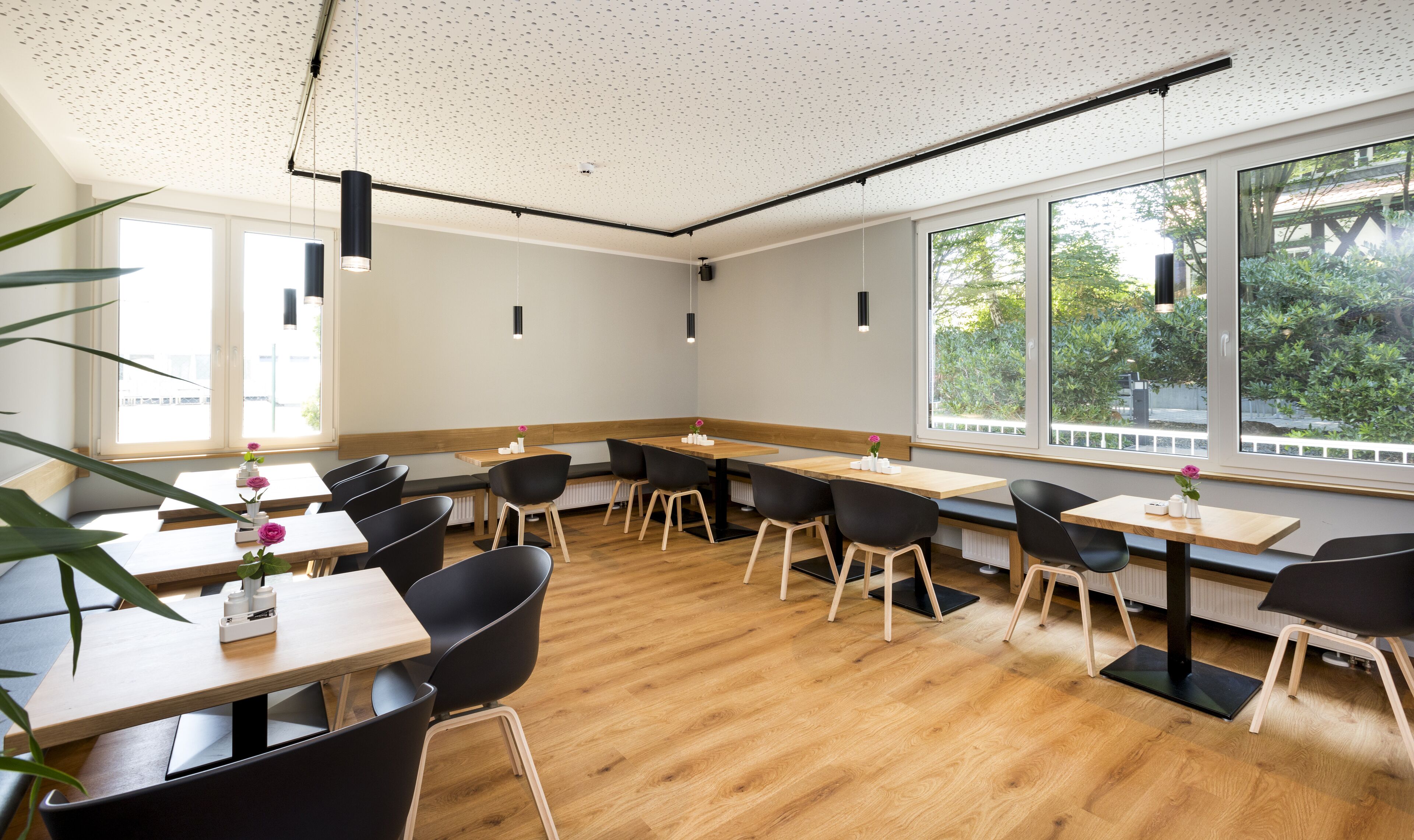 Uma sala de jantar moderna com mesas de madeira e cadeiras pretas, grandes janelas com vista para áreas verdes e iluminação pendente.
