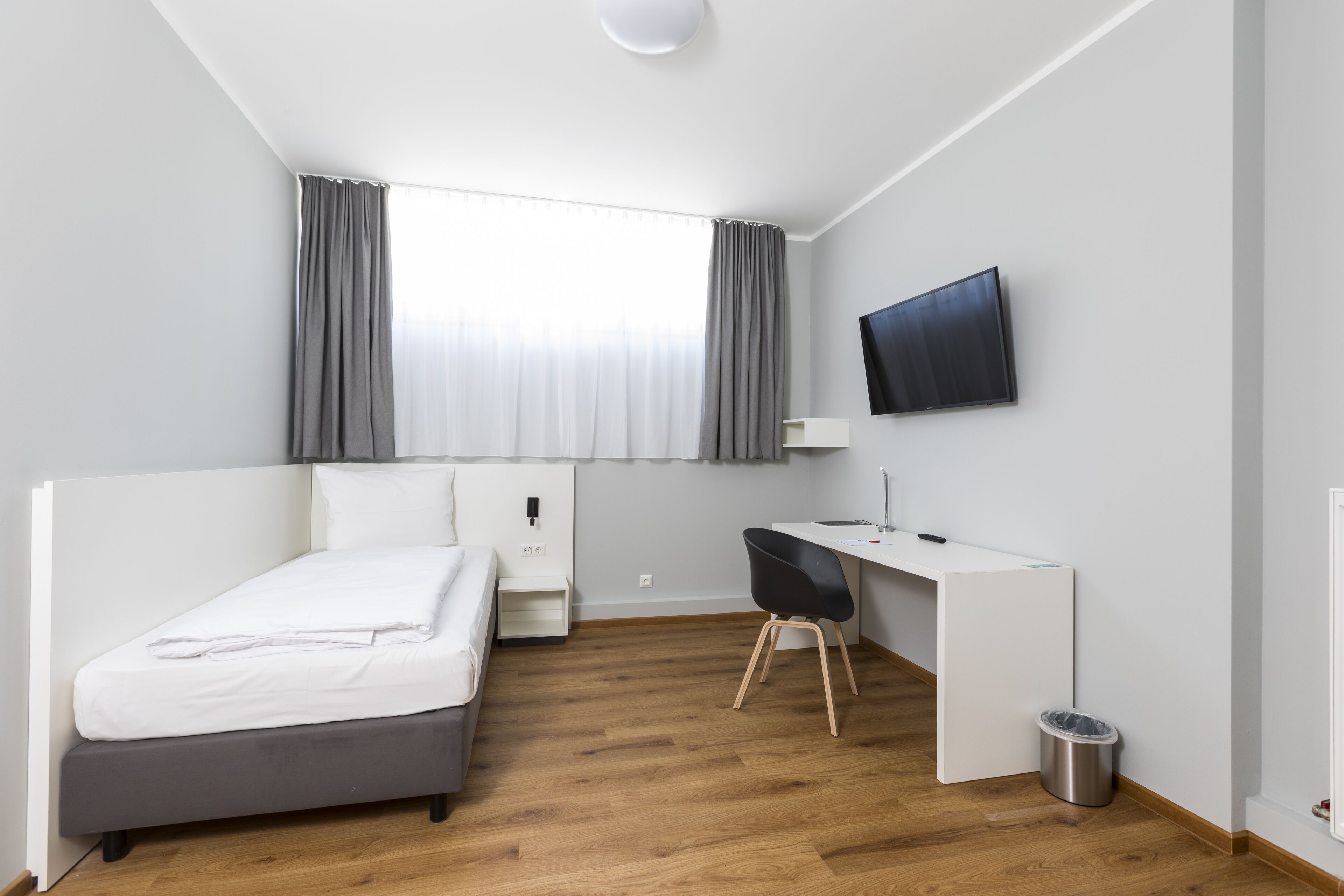 A modern hotel room with a single bed, a desk, a chair, a TV mounted on the wall, and a window with curtains.