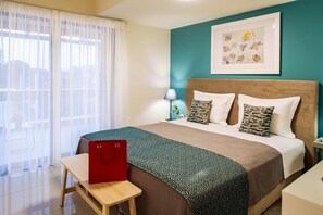 A hotel room with a bed, a nightstand, a lamp, a framed picture on the wall, and a window with sheer curtains. - Anemi Apartments I (Nafplio)