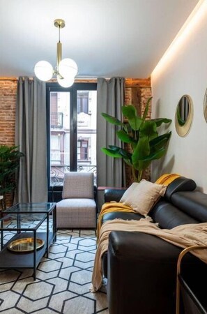 A modern living room with a black leather sofa, a glass coffee table, a white armchair, and a large green plant.