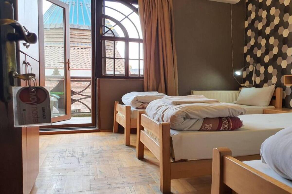 A room with two wooden beds, a window with curtains, a patterned wall, and a view of buildings outside.