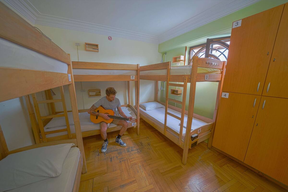 A bunk bed room with a person playing guitar, wooden bunk beds, a wooden wardrobe, and a window with a view.
