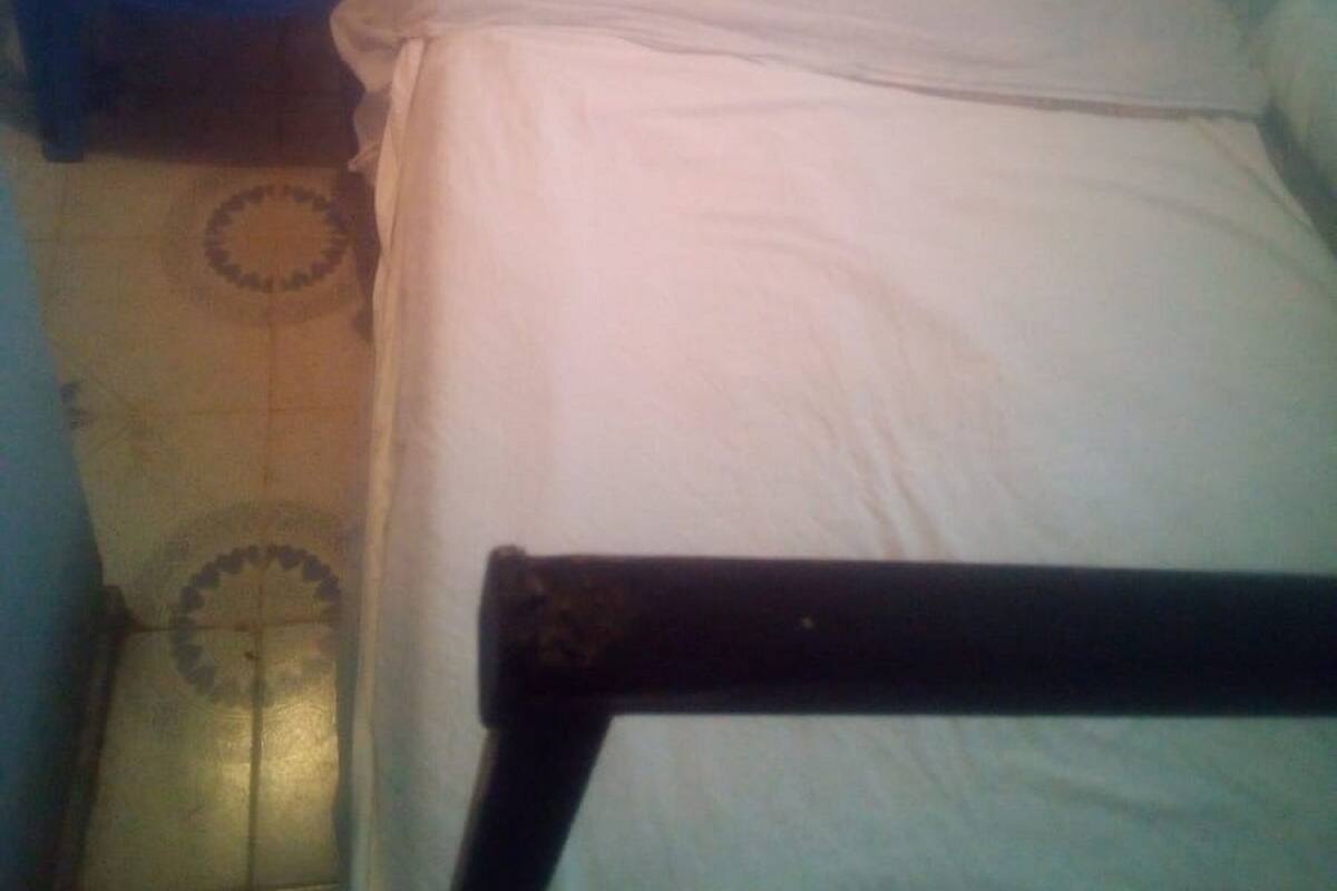A small, dimly lit room with a single bed, a blue plastic chair, and a tiled floor.