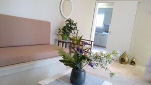 A living room with a sofa, a table with a vase of flowers, a mirror on the wall, and a view into the kitchen. - Palm tree house by the beach (Λουτράκι)