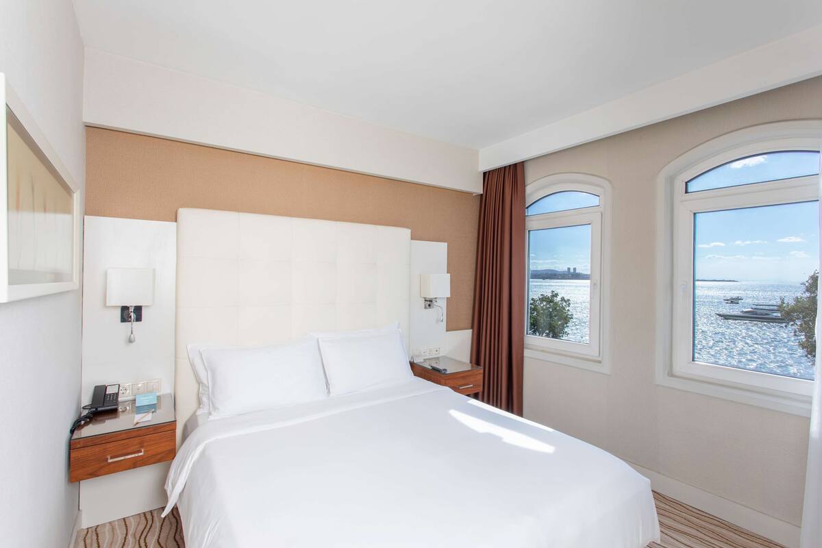 A hotel room with a large bed, a nightstand with a phone and lamp, and a window with a view of the water.