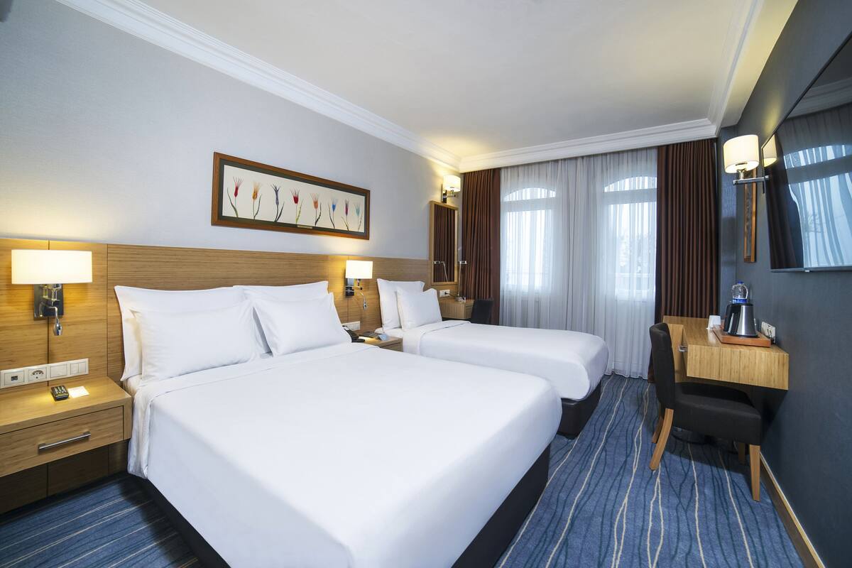 A hotel room with two beds, a desk, a chair, a lamp, a painting, and a window with curtains.