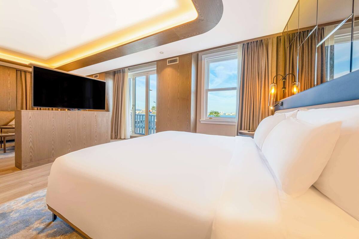A modern hotel room with a large bed, a flat-screen TV, a seating area, and large windows offering a view.