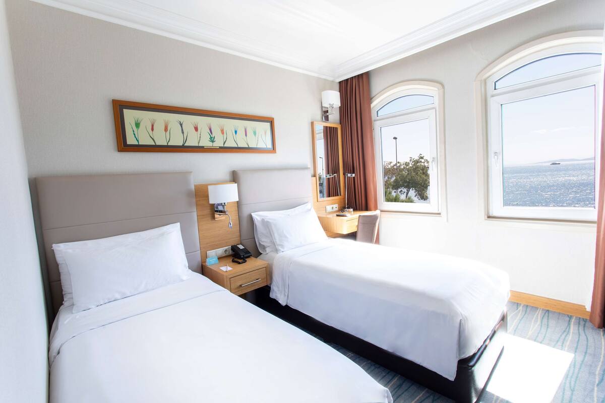 A hotel room with two beds, a bedside table with a lamp, a small desk, and a window with a view of the sea.