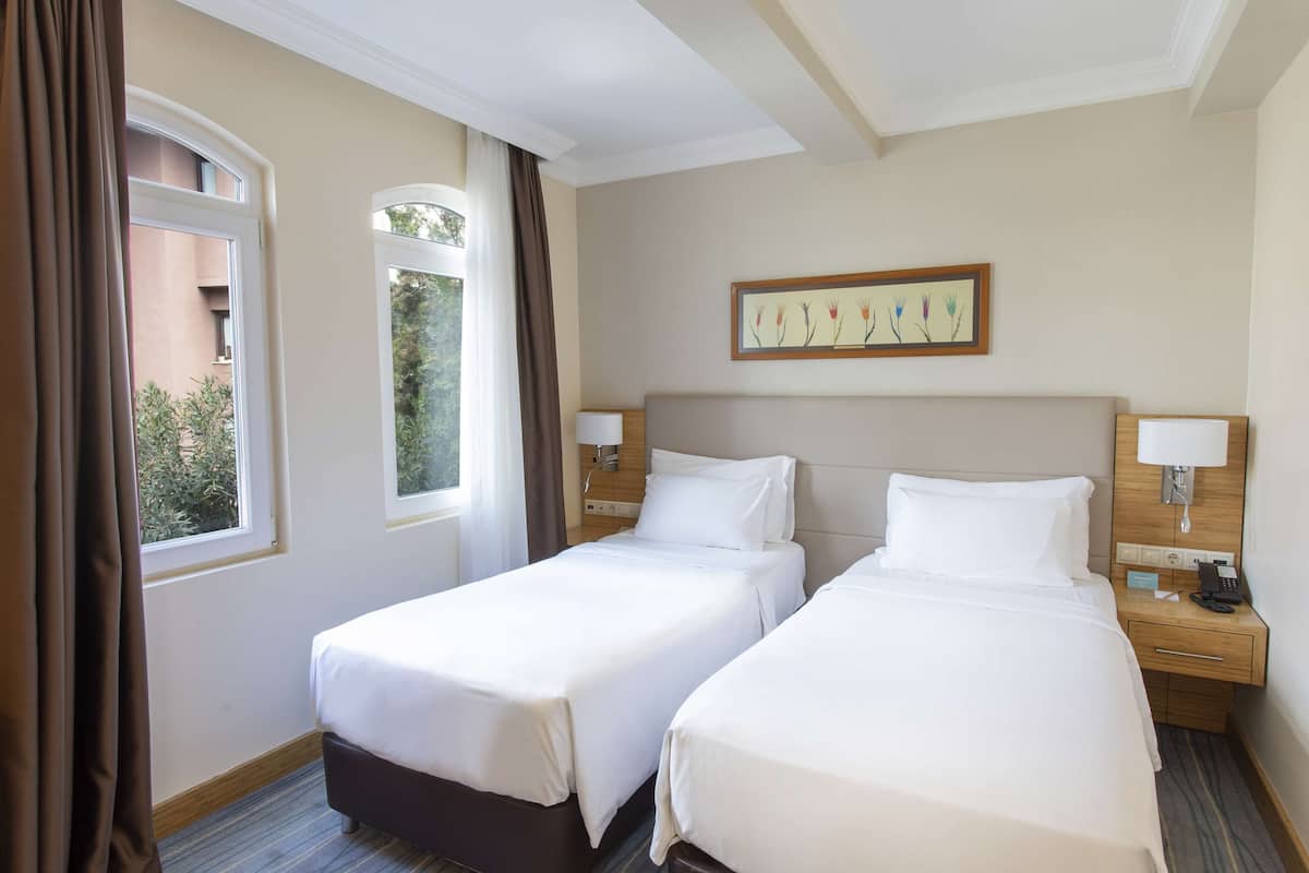 A hotel room with two single beds, a window with curtains, a wall-mounted artwork, and a bedside table with a lamp.