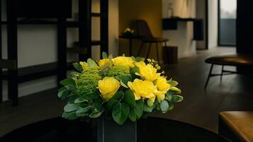 A modern living room with a dark wooden floor, a round black table with a square vase of yellow flowers, and a large window in the background.