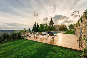 Um deck de madeira com espreguiçadeiras, piscina e um muro de pedra com plantas trepadoras.