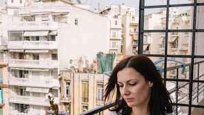 Una mujer sentada en una mesa de madera con una planta en maceta y una taza, con vista a una zona residencial con balcones y edificios.
