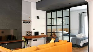 Una habitación de hotel moderna con una cama grande, un área de descanso con un sofá, un televisor y un panel de vidrio que separa el dormitorio de la sala de estar.