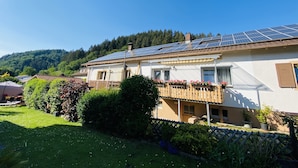 Une maison avec un toit équipé de panneaux solaires, un jardin clôturé et une vue sur les montagnes en arrière-plan.