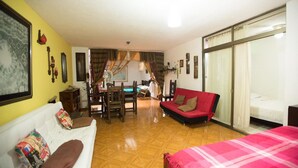 A room with a wooden floor, a red sofa, a white couch, and a bed with a pink blanket. There is a dining area with a table and chairs, and a window with curtains. - Charming 1-bed Apartment in Armenia (Armenia)