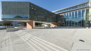 En moderne bygning med en stor glasfacade, stenelementer og en rummelig plads med trapper op til indgangen.