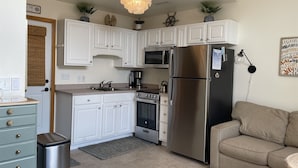 A compact kitchen with white cabinetry, a stainless steel refrigerator, a microwave, a stove, and a sink. There is a small trash bin and a beige sofa in the living area. - 38 Piece Of Heaven Studio Bedroom Condo by RedAwning (Hatteras)