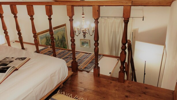 Une chambre avec un escalier en bois, un lit recouvert d’un drap blanc, un tableau accroché au mur et un lustre.