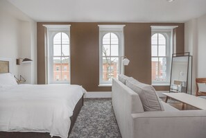 A hotel room with a bed, a sofa, a desk, and large windows offering a view of buildings outside.