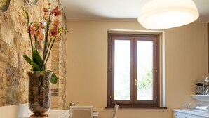 A kitchen with white cabinets, a dining chair, a window with wooden frames, and a hanging light fixture. - La Dimora di Francesco (Assisi)