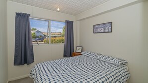 A bedroom with a large bed, a window with curtains, a framed picture on the wall, and a ceiling light. - The Black Shack - Cooks Beach Holiday Home (Cooks Beach)