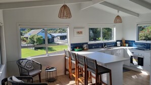 A modern kitchen with a large island, bar stools, and a view of the backyard. - The Black Shack - Cooks Beach Holiday Home (Cooks Beach)