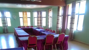 Un salón de conferencias con mesas y sillas, un ventilador de techo y ventanas grandes con cortinas.