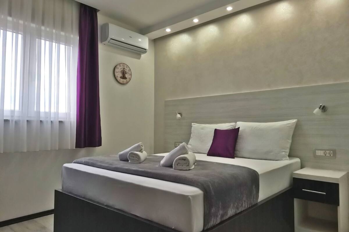 Une chambre d’hôtel moderne avec un grand lit, une table de chevet, une commode, un appareil de climatisation et une horloge murale.