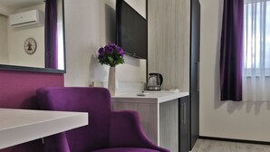 Una habitación moderna con un sillón púrpura, una mesa blanca, un jarrón con flores moradas, un televisor de pantalla plana y una cafetera.