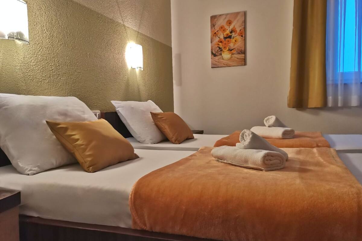 Une chambre d’hôtel avec deux lits, chacun agrémenté de draps blancs, d’oreillers marron et blancs, et d’une couverture orange. Un tableau orne le mur et une fenêtre est recouverte de rideaux.