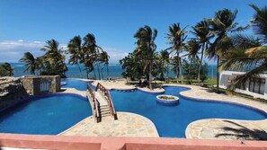 A pool area with a view of the ocean, surrounded by palm trees and a stone pathway leading to a building. - KimKev Smart Apartment (Shanzu)