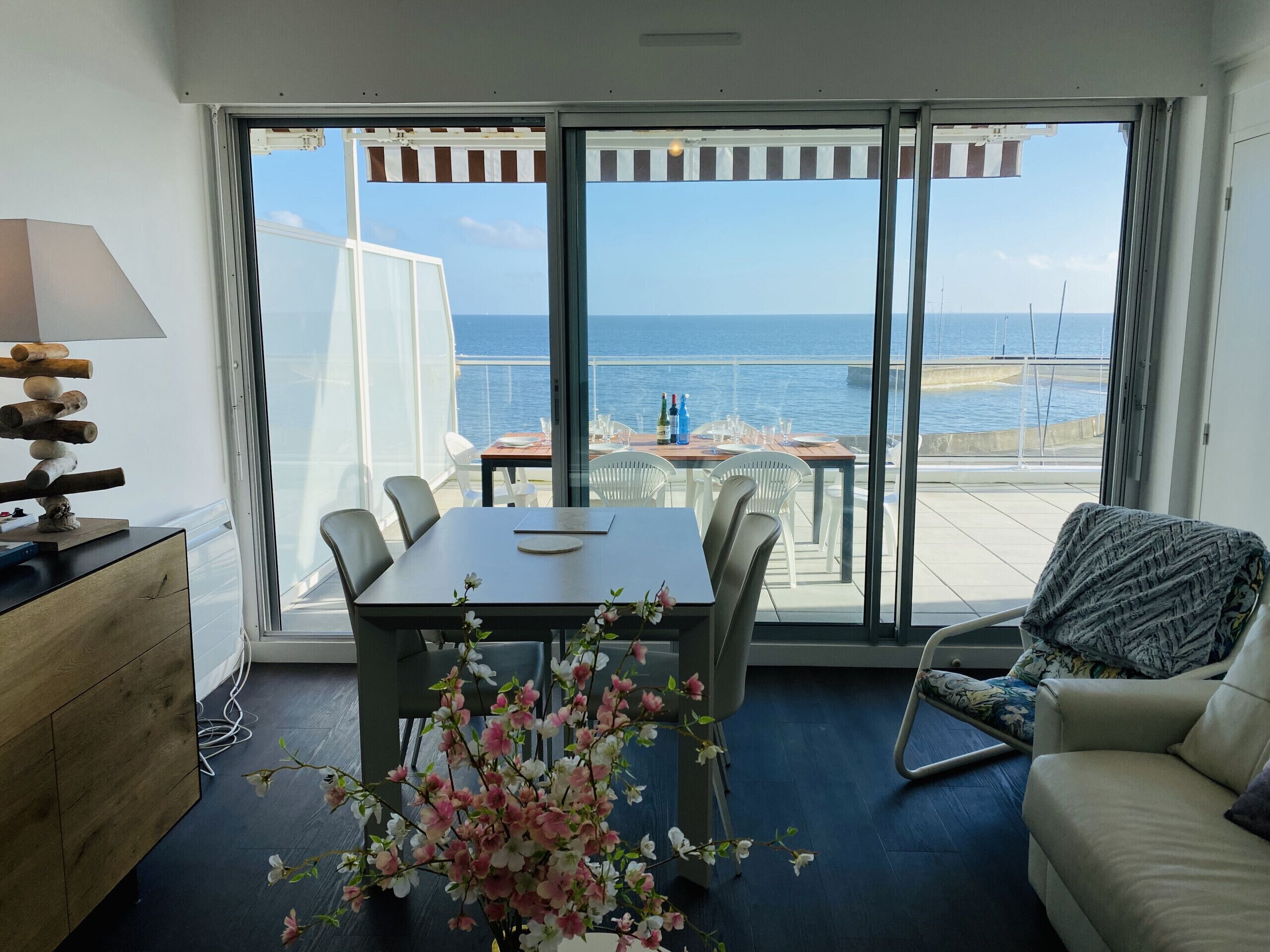 Ein Zimmer mit großem Fenster und Meerblick, einem für zwei Personen gedeckten Esstisch, einer Couch und einem Holzkleiderschrank.