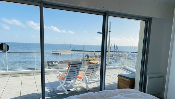 Ein Balkon mit einem Bett, zwei gestreiften Stühlen und Blick auf das Meer und Boote.