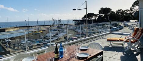 A terrace with a table set for dining, overlooking a marina with boats and a clear sky.