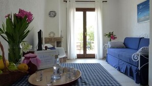 Sala de estar com chão xadrez, sofá azul, mesa de jantar com bandeja e porta para varanda.