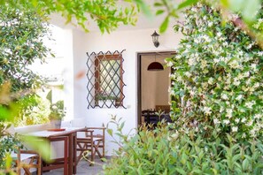 A patio area with a wooden table and chairs, a window with a decorative grille, and a potted plant. - At Lena's (Kassandra)
