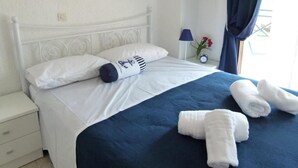 Uma cama bem arrumada com roupa de cama branca e detalhes em azul escuro, uma mesa de cabeceira branca com abajur e uma janela com cortinas azuis.