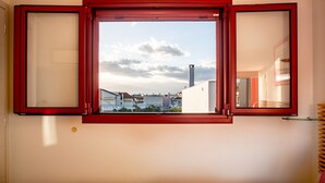 A room with a large window featuring a red frame, a view of buildings and a chimney outside. - Anemi  Apartments  II (Nafplio)