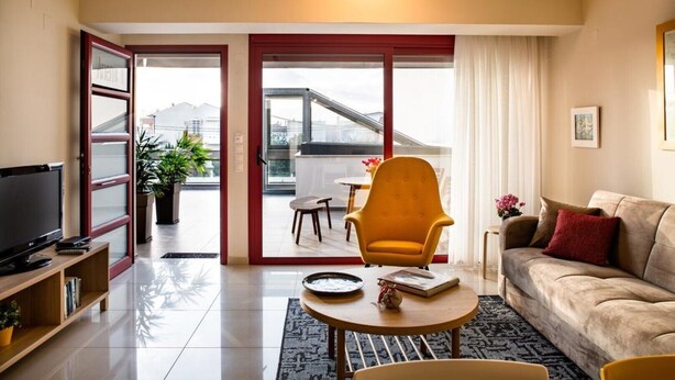 A modern living room with a sofa, a yellow armchair, a coffee table, and a TV. There is a balcony with a table and chairs, and a view of buildings. - Anemi  Apartments  II (Nafplio)