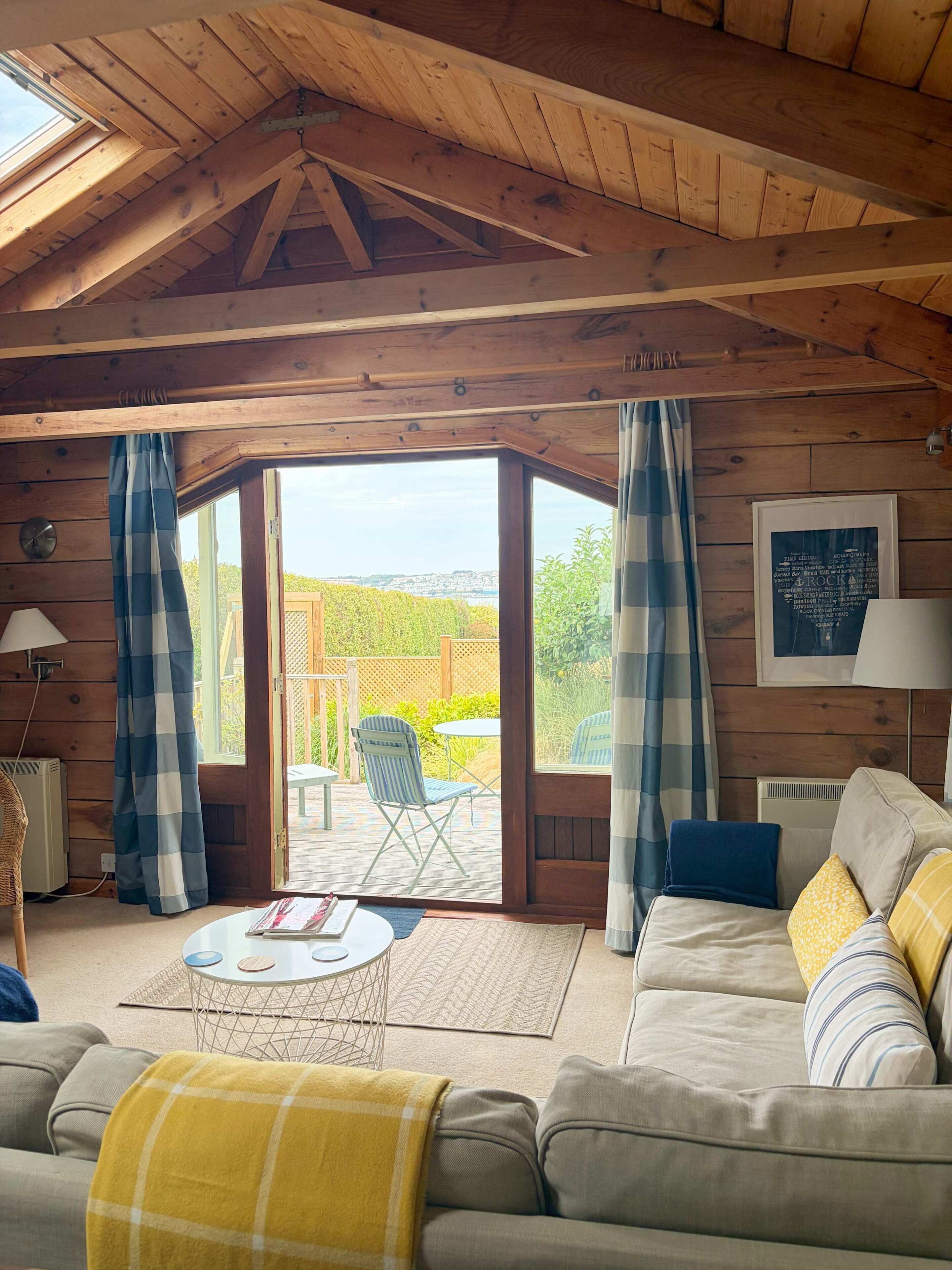 Ein gemütliches Zimmer mit Holzboden, einer Couch mit Kissen, einem Tisch und Blick durch eine Glastür auf einen Außenbereich mit Zaun und Grünpflanzen.