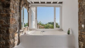 A bathroom with a large bathtub, a window, and a stone wall. - Villa Princess by Thea-concept (Mykonos)
