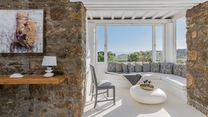 A cozy room with a stone wall, a wooden shelf with decorative items, a comfortable seating area with a sofa and cushions, and a large window offering a scenic view. - Villa Princess by Thea-concept (Mykonos)
