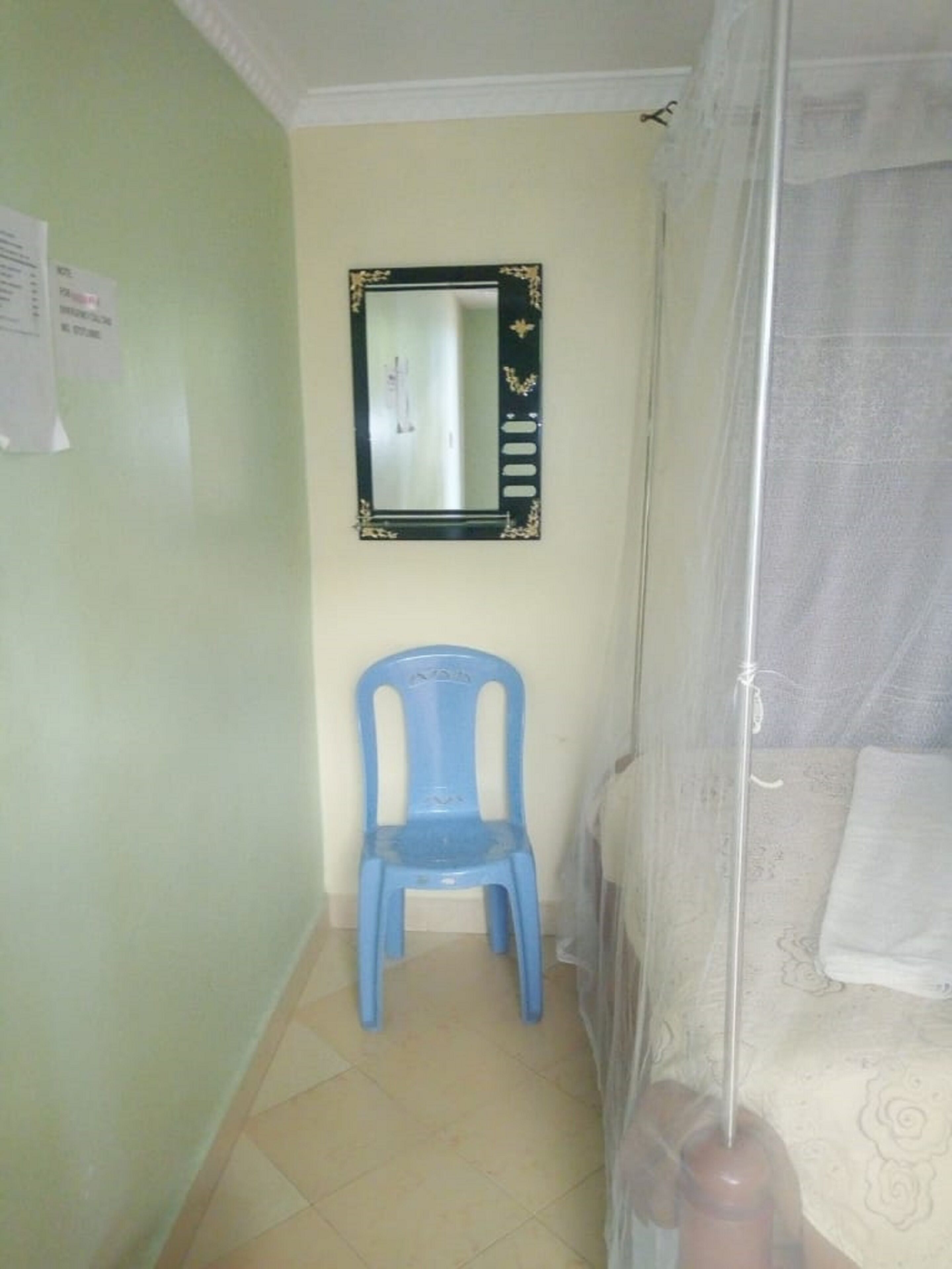 Una habitació petita amb una cadira de plàstic blau, un llit amb edredó blanc i un mirall fixat a la paret.