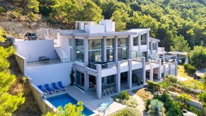 Une maison moderne à plusieurs étages, dotée d’une piscine, d’un espace de détente extérieur et offrant une vue sur une colline boisée.