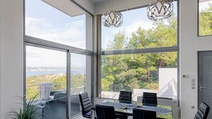 Un espace de repas moderne avec une table et des chaises en verre, de grandes fenêtres donnant sur l’extérieur et un luminaire suspendu.