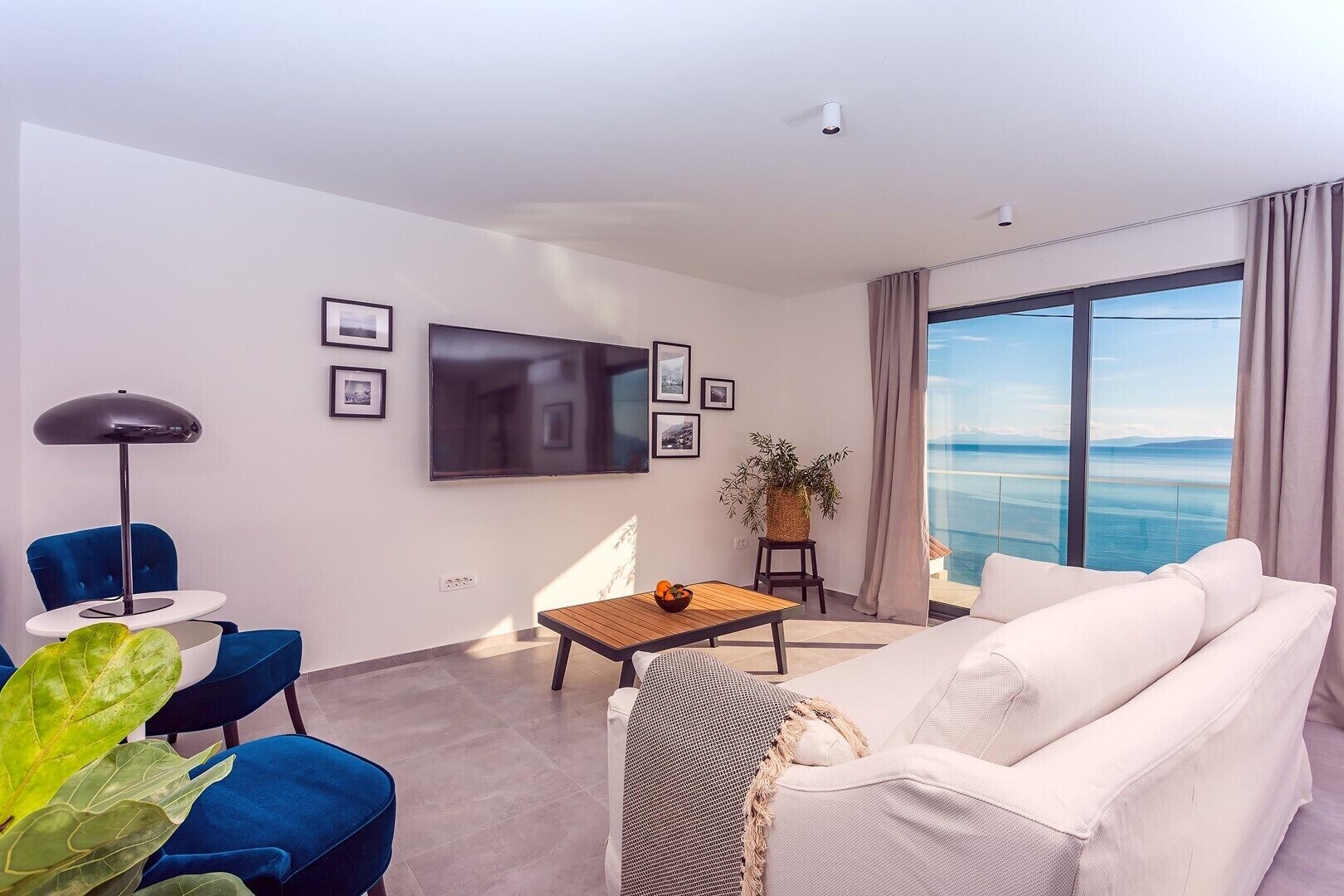 A modern living room with a large flat-screen TV, a blue chair, a wooden coffee table, and a white sofa.