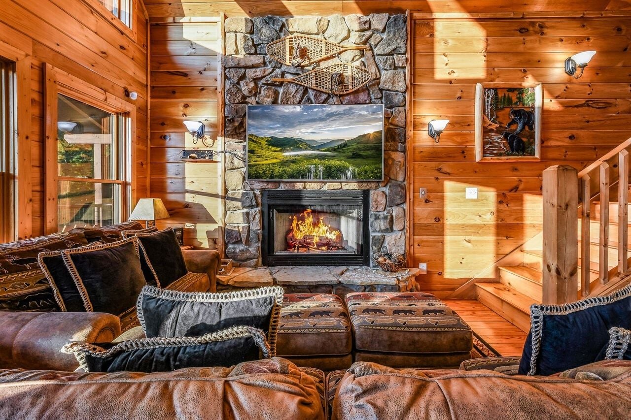 A cozy cabin interior with a fireplace, wooden walls, a stone fireplace surround, and a large window.